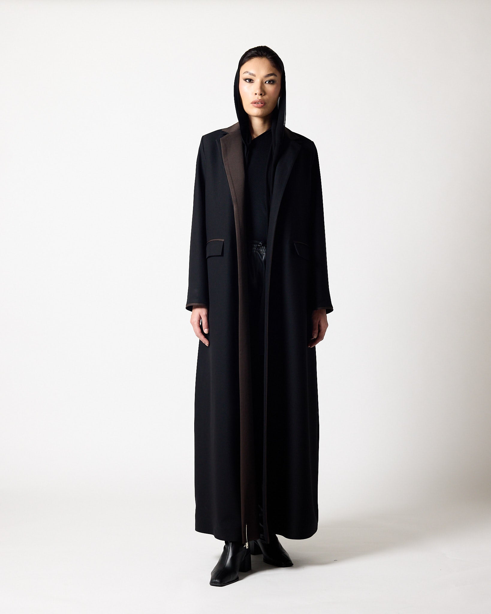 A person viewed from the back wearing a long black abaya or modest robe with a hood, standing against a plain light gray studio background.A model stands in a studio wearing a long, floor-length dark blue-gray abaya with a hood and a contrasting vertical front panel, paired with a black top and black shoes. A model wearing a long, dark abaya with a high collar and front slit, styled over a black top and black boots, standing against a light gray studio background.عبايات في قطر (Abayas in Qatar)محلات عبايات 
